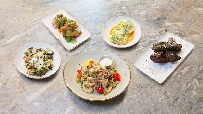 Aerial view of a stone table displaying five plated dishes. They include fried appetizers on white rectangular plates, a salad with mixed greens and cherry tomatoes in a round bowl, meat topped with sauce on a white plate, and garnished, sliced vegetables in a small bowl—the best Sunset Dining Maui offers.