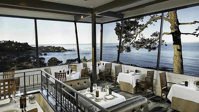 A restaurant with large windows overlooking a scenic coastal view on the Monterey Peninsula. Tables are set with white tablecloths, wooden chairs, glassware, and table settings. Outside, there are trees, cliffs, and an expansive ocean view under a clear sky—truly the best restaurant view around.