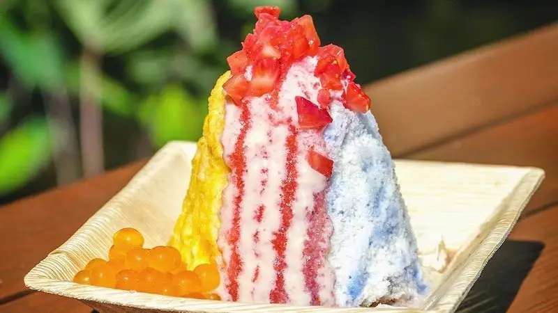 A colorful shaved ice dessert served on a square plate. The best shave ice in Waikiki features layers of yellow, red, and blue, topped with chopped strawberries and accompanied by a side of orange boba pearls. The background shows a blurred green scenery.