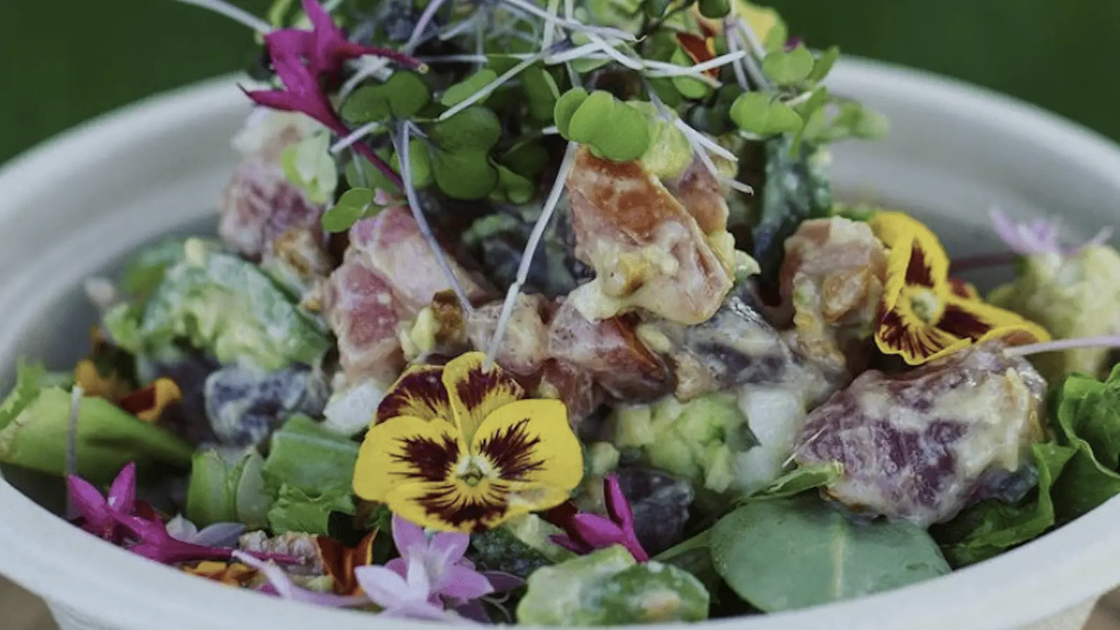 A colorful salad features diced vegetables, leafy greens, and edible flowers, topped with microgreens and drizzled with a creamy dressing, all reminiscent of the fresh flavors in the Best Poke Maui, presented in a white bowl.