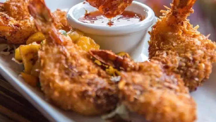 Close-up of a plate of golden-fried shrimp with a crispy coating, arranged around a small dish of red dipping sauce. The shrimp are accompanied by pieces of pineapple garnished with herbs, creating an appetizing and colorful presentation—perfect for the best happy hour in Kauai.
