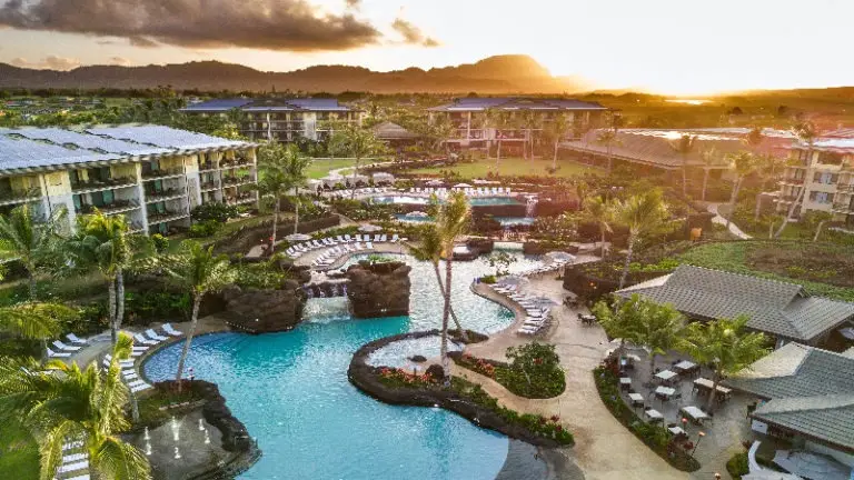 An aerial view of the Poipu resort at sunset, showcasing multiple pools surrounded by lounge chairs and palm trees. Buildings with balconies can be seen around the pool area. The backdrop features lush greenery and mountains under a partly cloudy sky, perfectly capturing the essence of Koloa Landing Resort.
