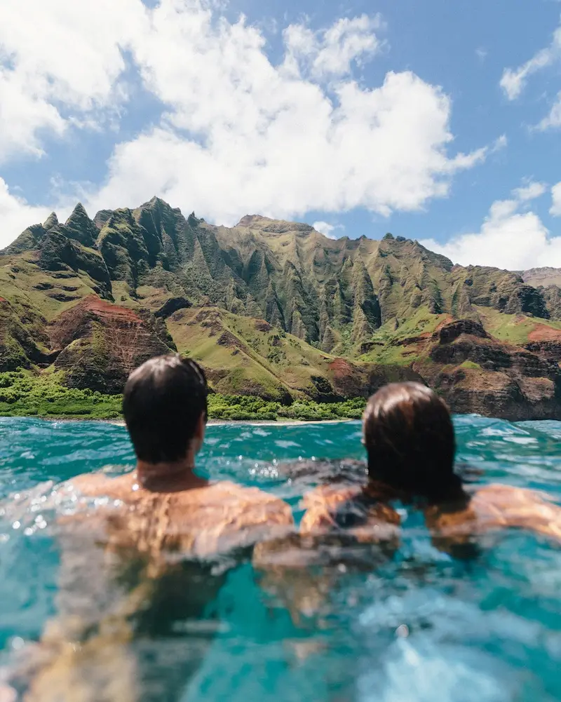 A man and a woman swimming in clear blue water, facing a lush, green, and rocky mountainous shoreline under a bright blue sky with scattered clouds.