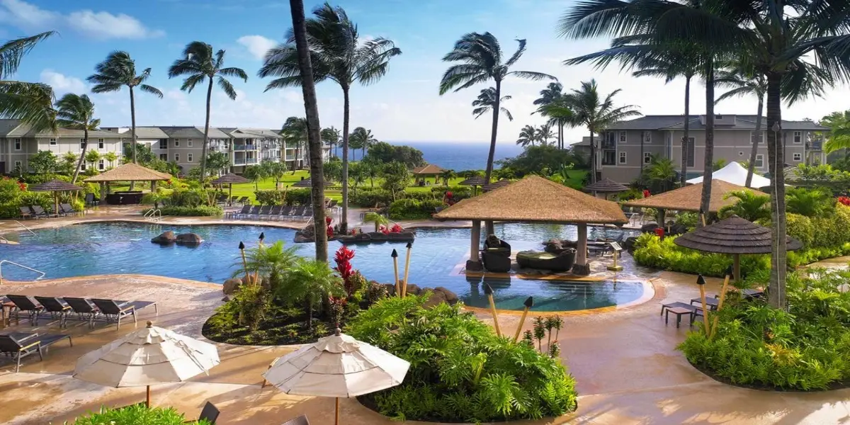 A tropical luxury resort, The Westin Princeville Ocean Resort Villas, features a large swimming pool surrounded by palm trees, sun loungers, and thatched-roof cabanas. The area is lush with green foliage and faces the ocean. Buildings with balconies are in the background, and the sky is clear with scattered clouds.