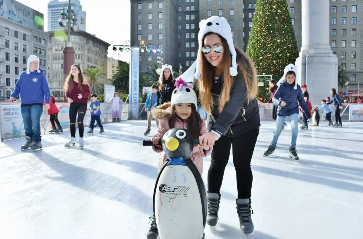 A woman and a child, both wearing animal-themed hats, are ice skating together at one of the best things to do in the Bay Area this November. The child is holding onto a penguin skate aid while the woman supports her from behind. Other skaters and a large decorated Christmas tree are visible in the background.