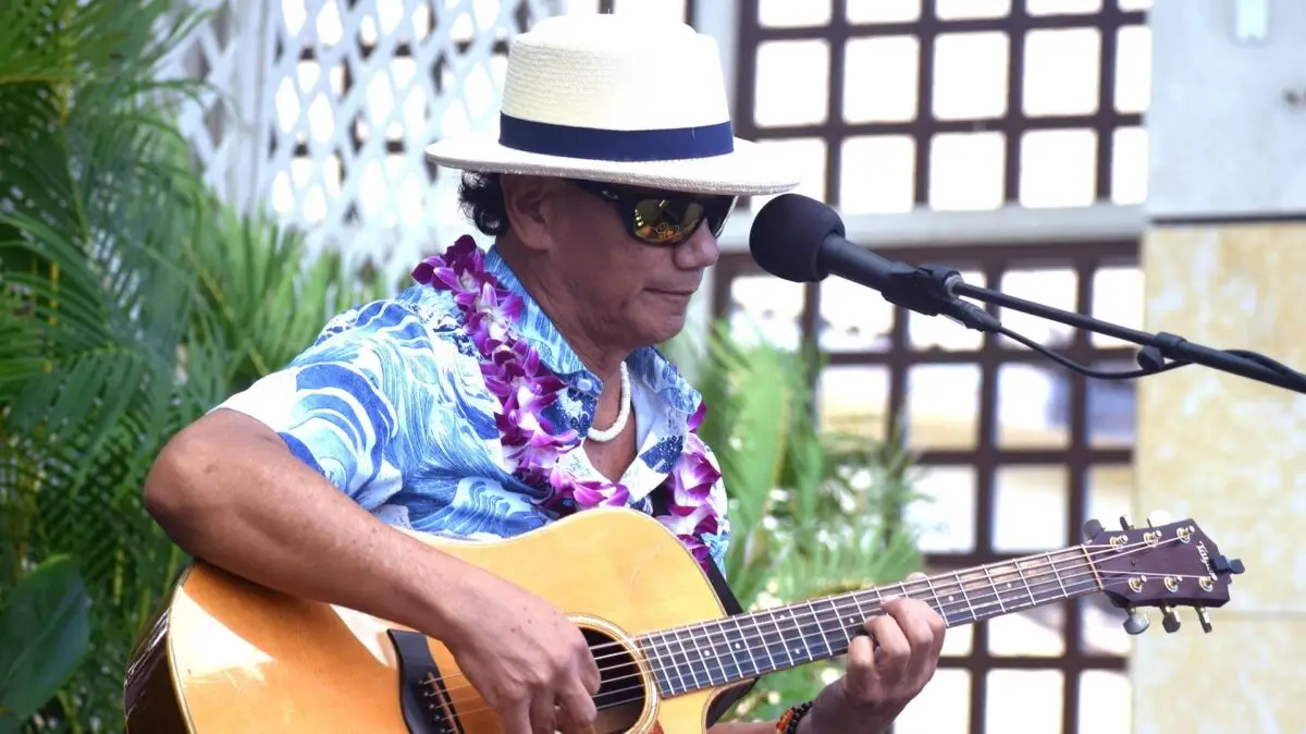 A man wearing a beige hat, sunglasses, a colorful Hawaiian shirt, and a flower lei plays an acoustic guitar into a microphone at the Outrigger Hotel on the Big Island. The background features a wooden lattice and lush green foliage, suggesting an outdoor, tropical setting.