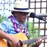 A man wearing a beige hat, sunglasses, a colorful Hawaiian shirt, and a flower lei plays an acoustic guitar into a microphone at the Outrigger Hotel on the Big Island. The background features a wooden lattice and lush green foliage, suggesting an outdoor, tropical setting.