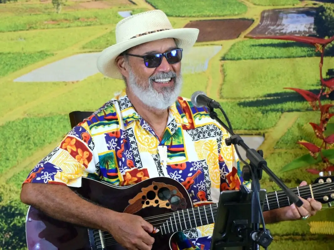 A man with a white beard and sunglasses is playing an acoustic guitar. Wearing a colorful Hawaiian shirt and straw hat, he sings into a microphone against the lush, green landscape of fields and trees. It's a perfect day at the Outrigger Hotel Big Island.