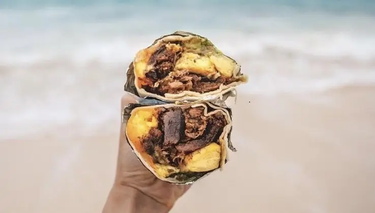 A hand holding the best breakfast burrito on the North Shore, cut in half, revealing scrambled eggs, meat, and other fillings inside. The background is a blurred view of a sandy beach and ocean waves.