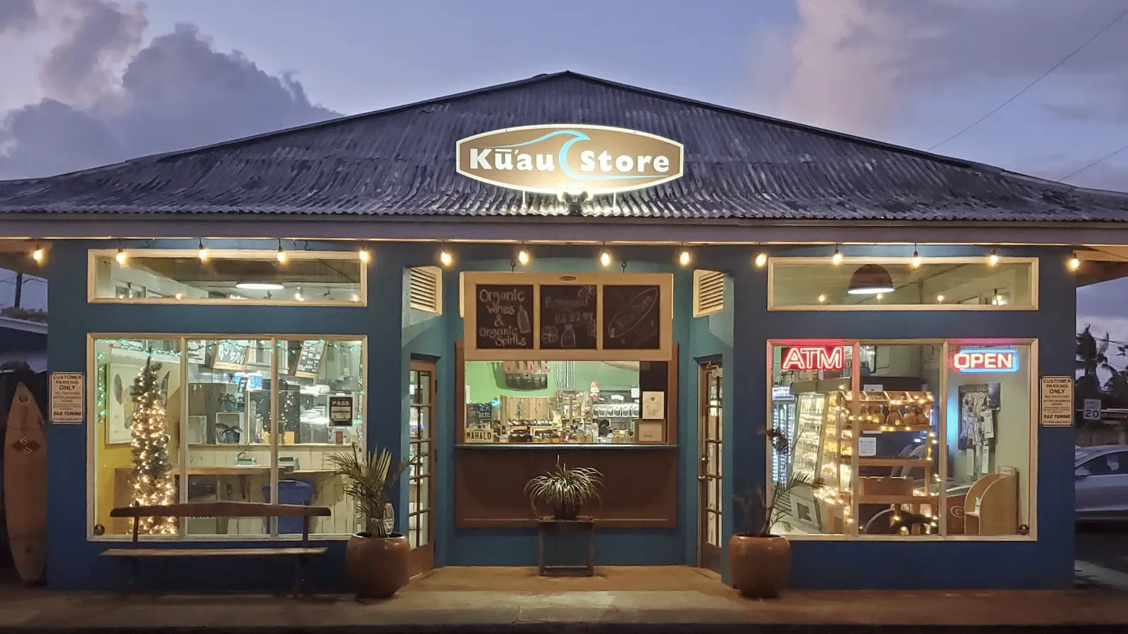 A small, blue-painted store named "Kūau Store" is lit up at twilight. The facade displays a surfboard to the left, festive lights inside, and a chalkboard menu above the entrance. Signs include "ATM" and "OPEN," indicating that the store, known for the Best Poke Maui, is open for business.