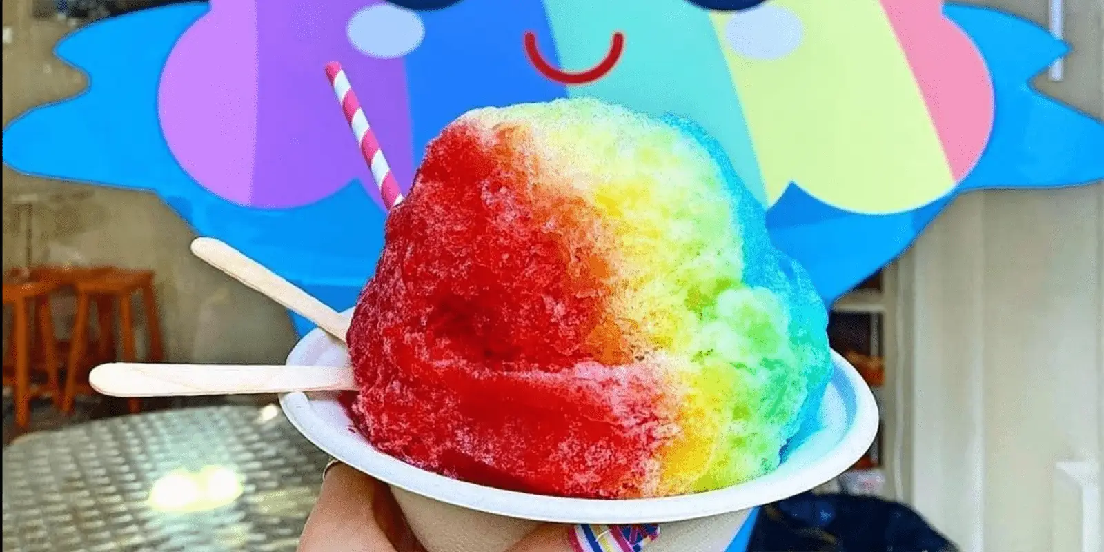 A colorful shaved ice treat in a white cup, featuring vibrant red, yellow, green, and blue sections. Served with a striped straw and two wooden spoons, it boasts the best shave ice in Waikiki. The background shows a cheerful, cartoon-like mural with a smiling face and rainbow colors.