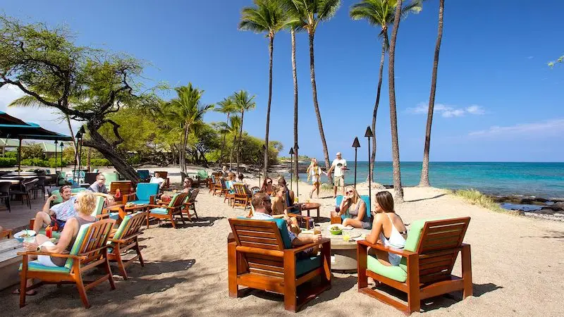 A group of people relaxes on wooden chairs at a sandy beachside cafe with food and drinks during the best happy hour on the Big Island. They are sitting under palm trees with the ocean in the background. The sky is clear and blue. Some people are standing and taking photos.