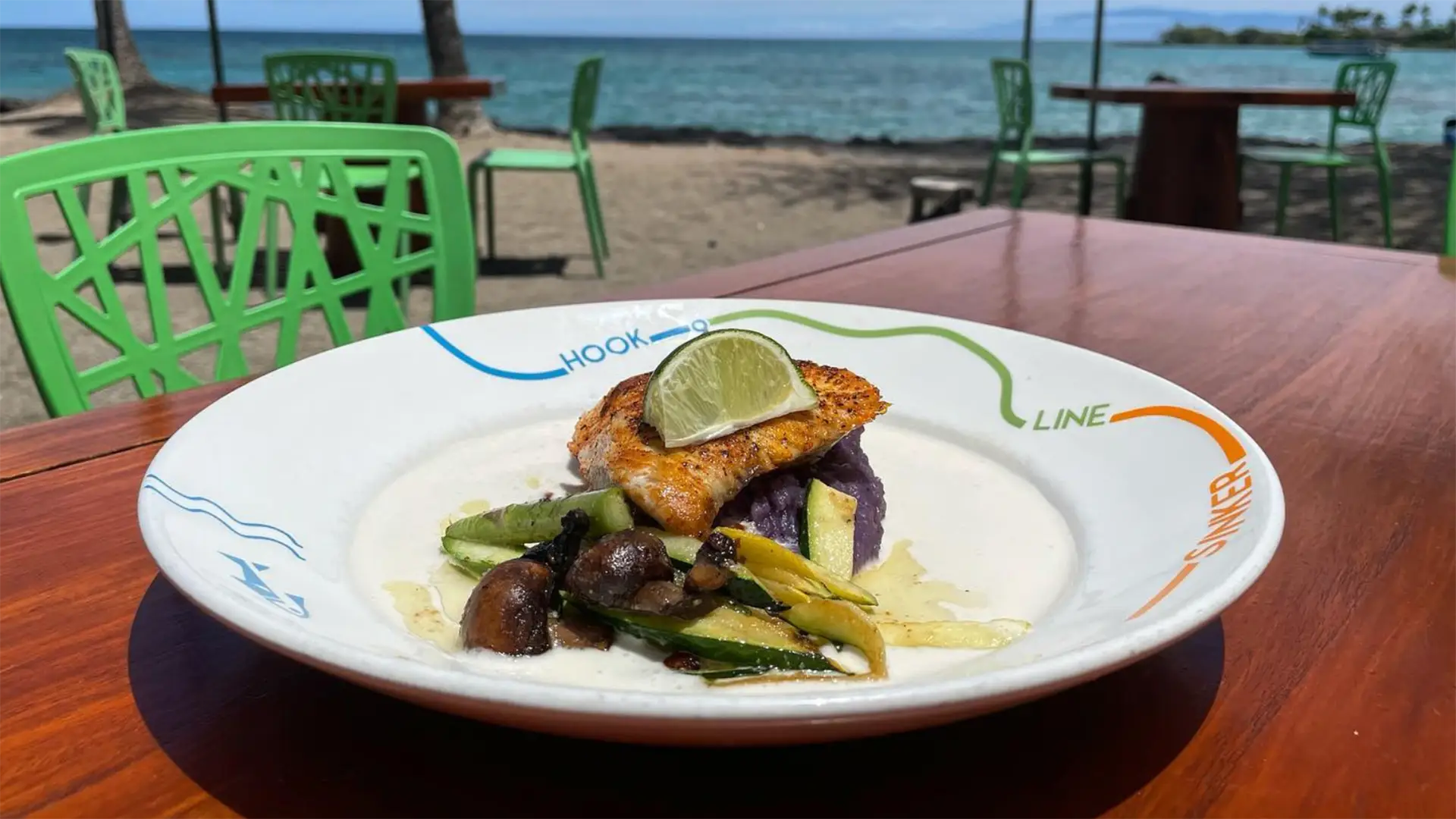 A plate of grilled fish garnished with a lime wedge, served with sautéed vegetables sits on a wooden table at an outdoor restaurant with green chairs. The backdrops are a sandy beach, palm trees, and the blue ocean under a clear sky—a perfect scene for enjoying the best Hawaiian food on the Big Island.