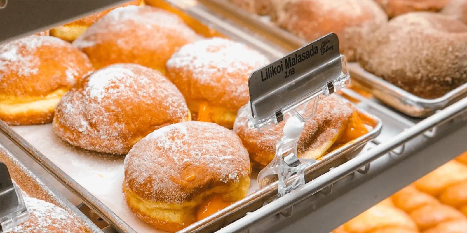 A display case filled with trays of Lilikoi Malasadas, round, fluffy doughnuts dusted with powdered sugar, some oozing with a creamy filling. A small sign labels the pastries with their name and price. These are renowned as the best malasadas in Honolulu.