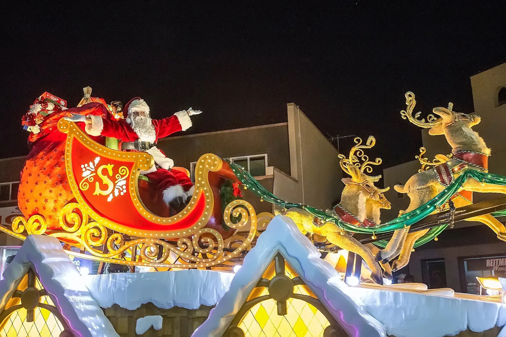 Santa Claus, sitting in a festive red sleigh filled with gifts, waves and points while being pulled by reindeer through a snowy, nighttime scene. Bright decorative elements adorn the sleigh, and the background shows buildings lit up by Christmas lights—one of the best things to do South Bay November!