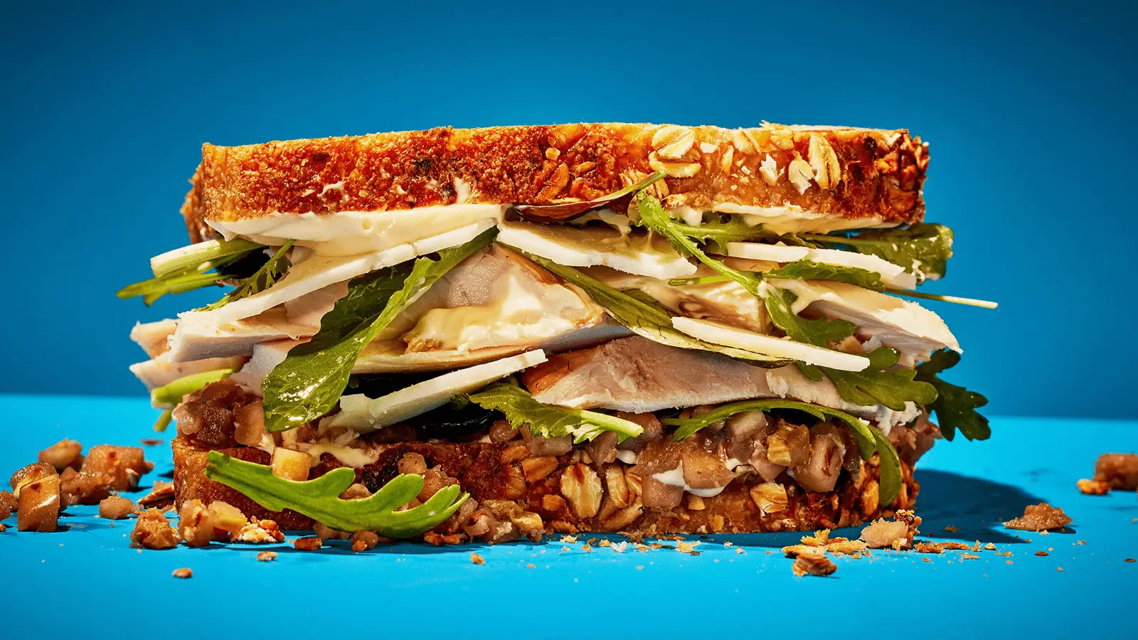 A close-up of a sandwich on multigrain bread, filled with slices of roasted turkey, leafy greens, cucumber slices, and a light spread of mustard or mayonnaise, all against a bright blue background. Crumbled bread and filling pieces are scattered around the base—truly the best lunch in Napa Valley.