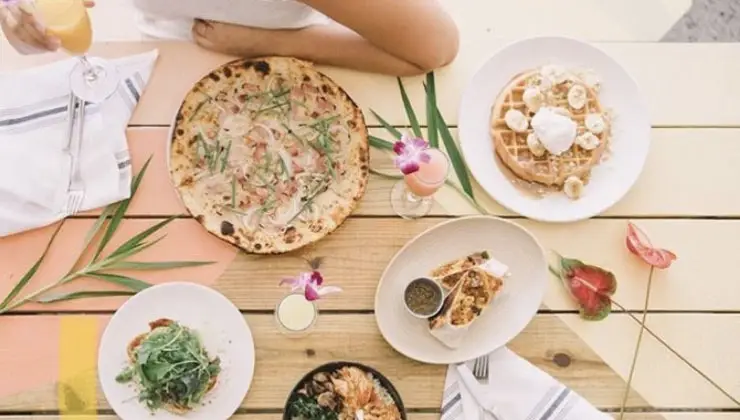 A table set with various foods, including a pizza topped with greens, a waffle with cream and banana slices, a salad, and other dishes. A person is seated with a drink in hand, enjoying the best happy hour Waikiki has to offer as the table is adorned with flowers and greenery.