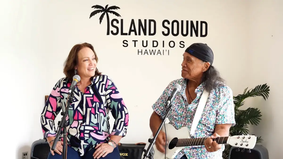 Two musicians perform in front of a wall with the text "Island Sound Studios Hawai'i" and a palm tree logo. One is seated and singing into a microphone, wearing a colorful patterned shirt. The other stands playing a white guitar, wearing a patterned shirt and headband, celebrating Henry Kapono on Make Music Day.