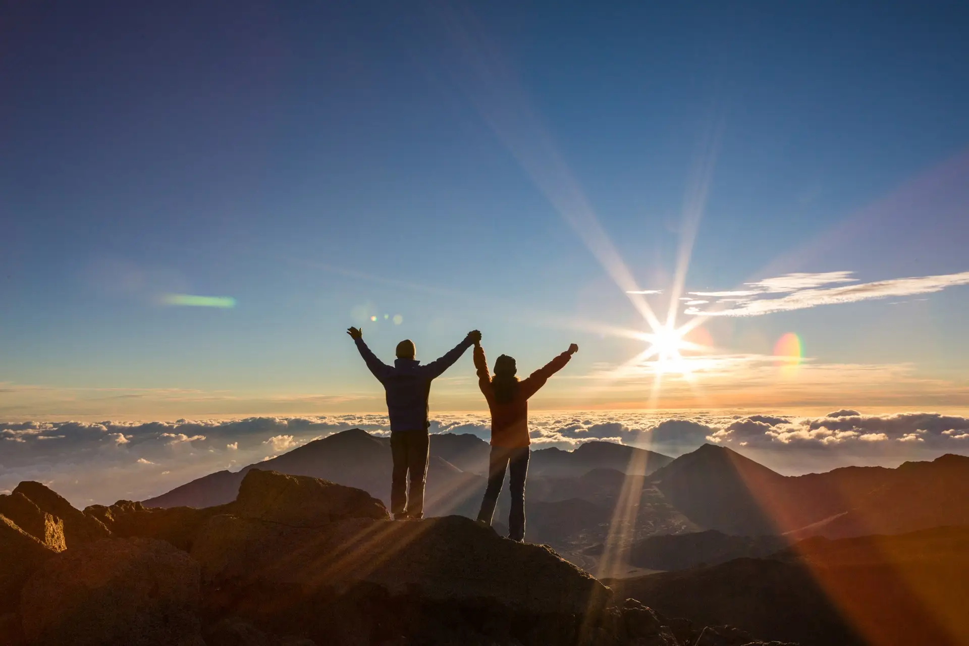 Two people stand on a mountain peak at sunrise, silhouetted against the sky. They hold hands and raise their arms in celebration. Below, layers of mountains and clouds stretch out, illuminated by the golden sunlight.