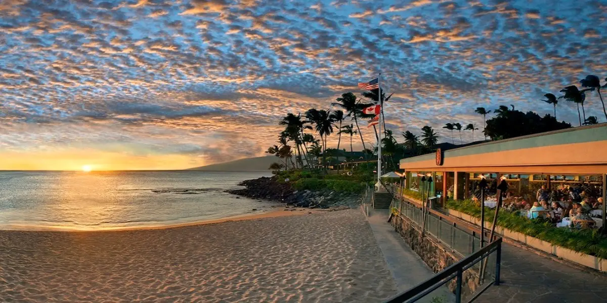 A scenic beach at sunset with golden clouds, gentle waves, and silhouettes of palm trees. A cozy restaurant to the right has outdoor seating, some patrons visible. American and other flags fly on poles near the building, enhancing the tranquil atmosphere characteristic of Napili Kai Resort.