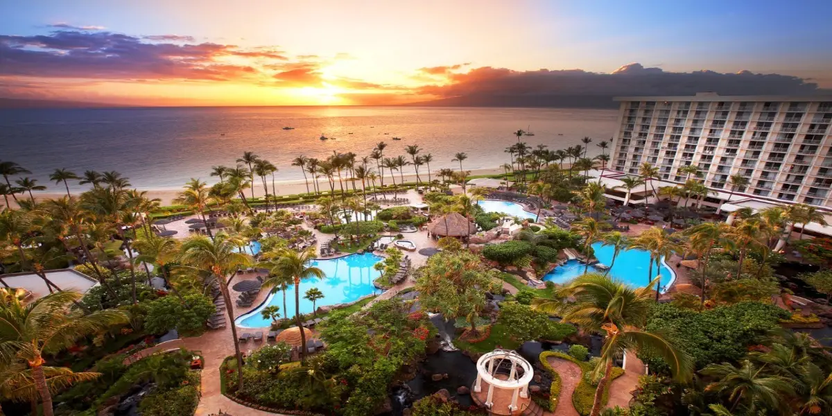 Aerial view of the Westin Kaanapali Resort at sunset featuring multiple pools, lush palm trees, and manicured gardens. A multi-story hotel building stands on the right, while the sun sets over the ocean in the background, casting vibrant colors across the sky.