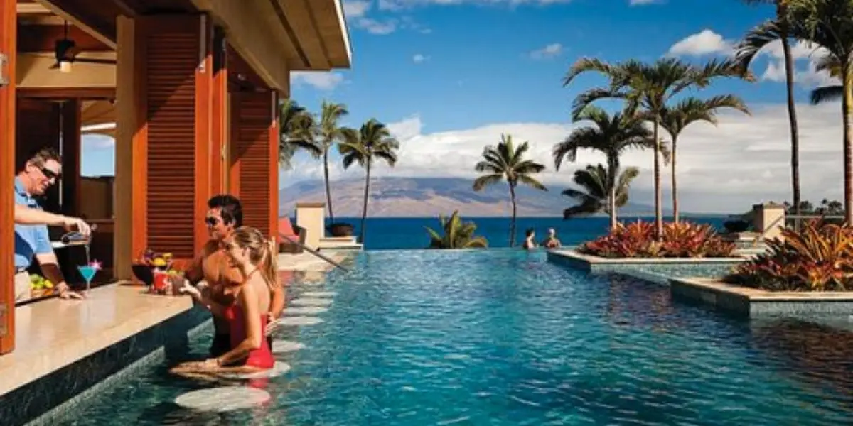 A luxurious pool scene at Four Seasons Maui with people enjoying a swim and drink. A man serves colorful drinks to two adults seated by a pool bar. The background features palm trees and an ocean view with a mountain in the distance under a partly cloudy sky.