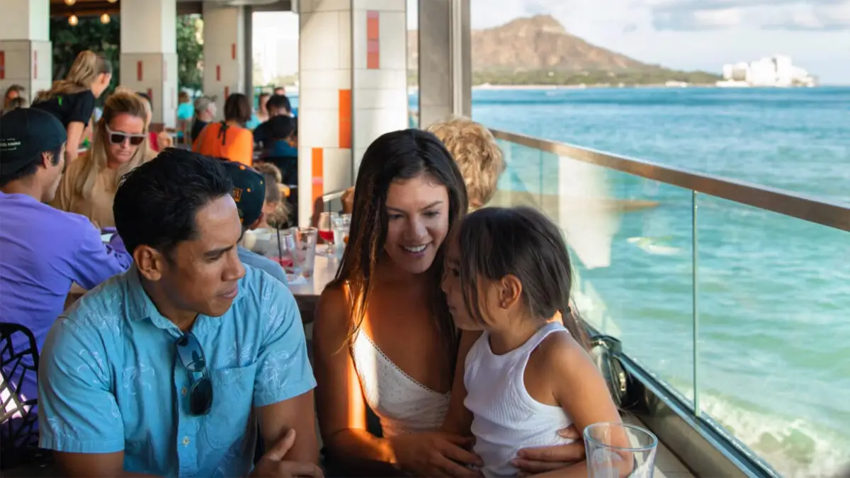 A family of three sits at a restaurant with ocean views. The man on the left wears a blue shirt, the woman in the center holds a child and smiles, and the child looks at the woman. Other diners are in the background, and a mountain is visible across the water.