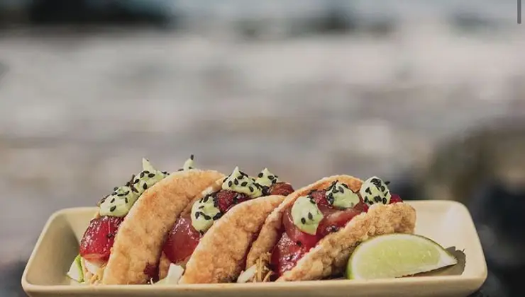 A close-up of three tacos filled with fish, vegetables, and toppings sits on a rectangular plate. The tacos are garnished with a light green sauce and seeds. A wedge of lime is placed on the side of the plate. The background appears to be a blurred outdoor setting, ideal for the best Happy Hour Kaanapali Maui offers.