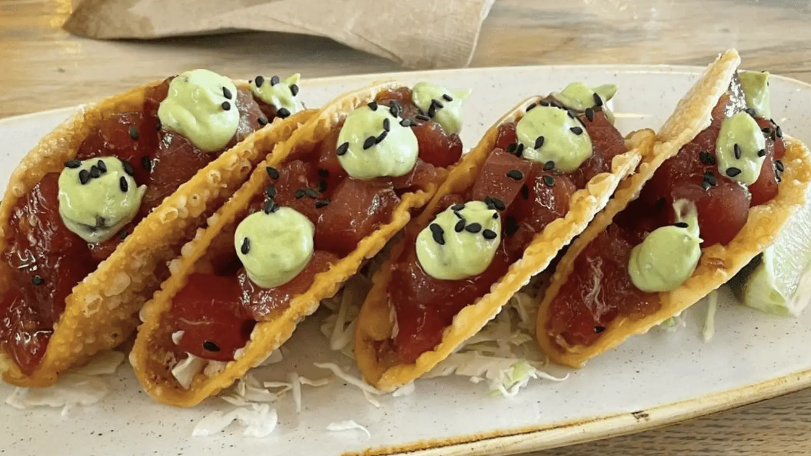 A plate of four crispy tacos filled with diced tuna, topped with green avocado sauce and sprinkled with black sesame seeds from Best Poke Maui. A wedge of lime is visible on the side. The tacos are presented on a white plate on a wooden table.