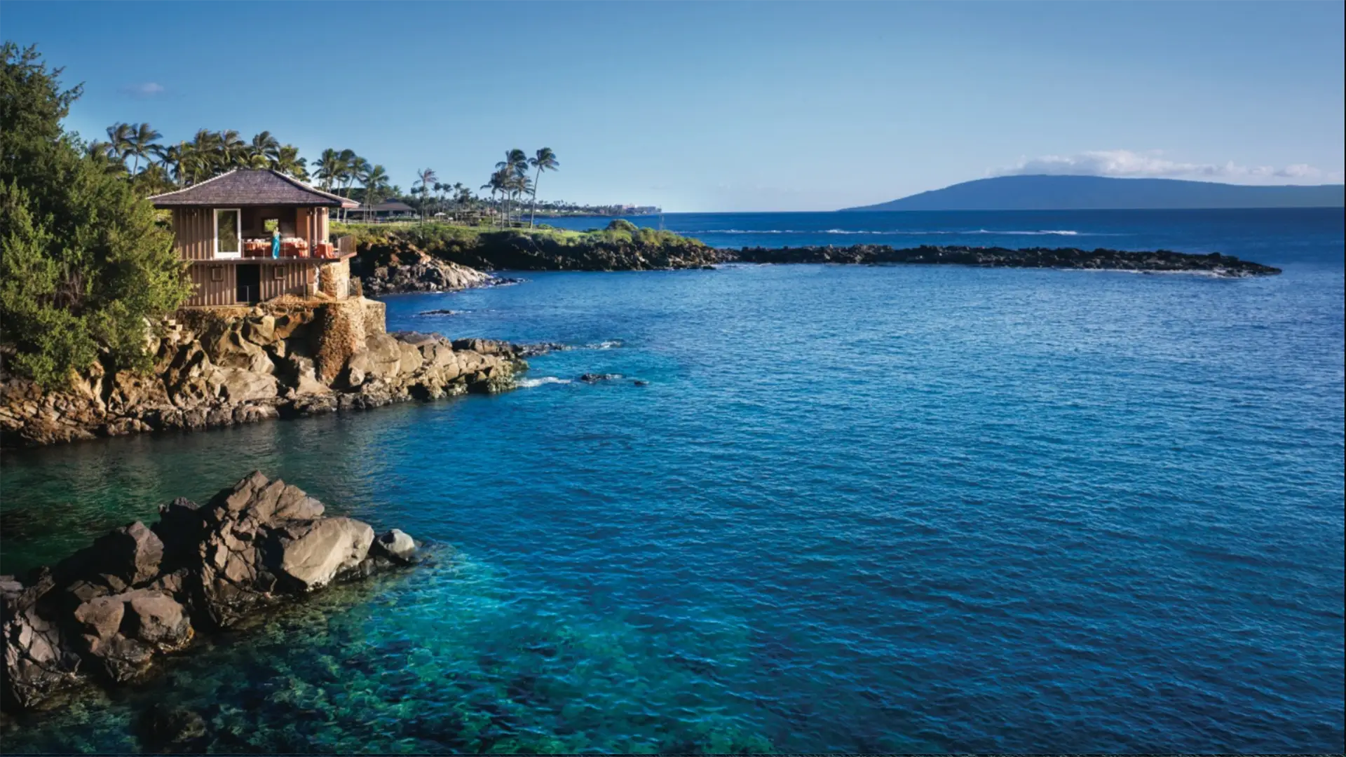 A picturesque seaside landscape shows a house perched on a rocky cliff overlooking a clear blue ocean. The water gently laps against the rocks and lush green vegetation surrounds the area, reminiscent of some of the best romantic hotels in Maui West. In the distance, a mountainous island rises under a clear, blue sky.