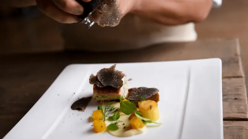 A chef grates truffles over an elegantly plated dish on a white rectangular plate. The dish features small cubes of what appears to be potato or polenta, garnished with fresh greens and drizzled sauce, creating a refined and artistic presentation—one of the best things to do in Napa Valley this January.