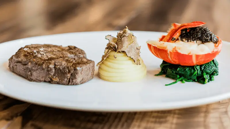A gourmet dish featuring a seared beef steak on the left, a mound of mashed potatoes topped with slices of truffle in the center, and a lobster tail filled with black caviar on a bed of greens on the right, all plated on a white dish—one of the best things to do in Napa Valley this January.