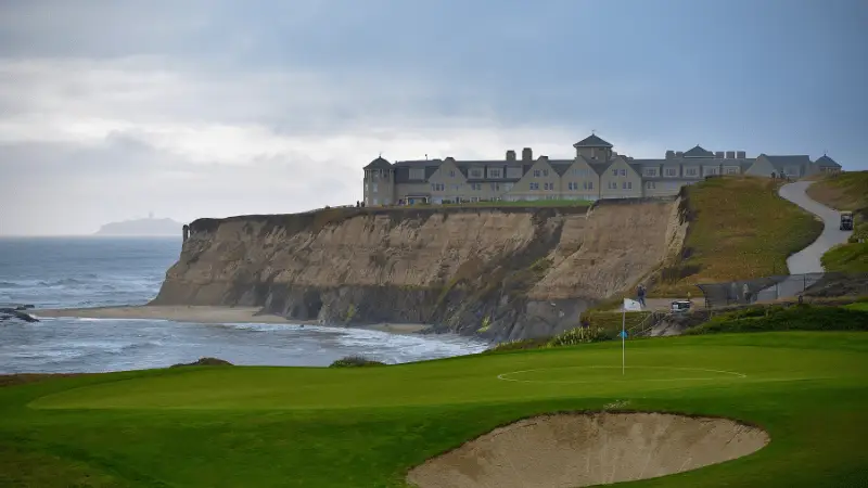 A luxury hotel in Half Moon Bay sits atop a rugged cliff overlooking the ocean, with waves crashing below. In the foreground, a manicured golf course with a green and sand trap is visible. A winding path leads from the golf course up to the hotel, offering the perfect coastal getaway. The sky is overcast.
