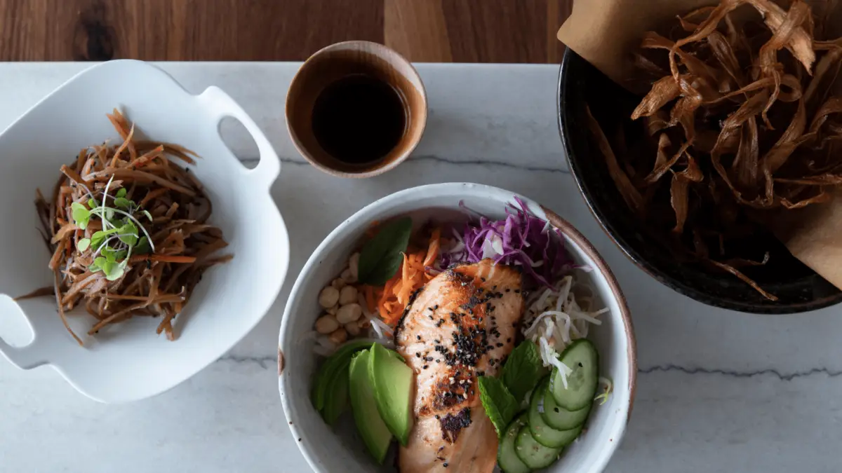 Top view of a meal comprising a white bowl filled with salmon, avocado slices, shredded carrots, red cabbage, cucumber slices, peanuts, and rice, garnished with black sesame seeds. Side dishes include a bowl of fried elements and a white tray of shredded vegetables—truly the Best Dinner on Maui.