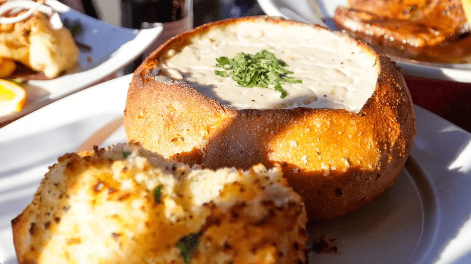 A bread bowl filled with the best clam chowder Monterey Peninsula has to offer, garnished with chopped herbs, sits on a white plate. A piece of garlic bread is placed in the foreground, with other dishes slightly blurred in the background. The scene is well-lit, highlighting the textures of the food.