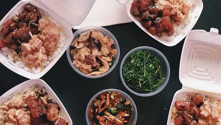 A variety of takeout dishes are arranged on a table. The selection includes the best poke Waikiki Oahu has to offer, with fresh raw fish, containers of mixed greens, and shredded vegetables. The food is served in white foam containers with white disposable napkins neatly placed in the center.