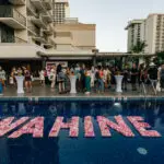 A diverse group of people gathers by a pool at one of the luxurious Outrigger Hotels on Oahu, socializing and enjoying an outdoor event. The pool features a floating decoration with "WAHINE" spelled out in pink flowers, while surrounding buildings and palm trees create a modern, tropical setting.