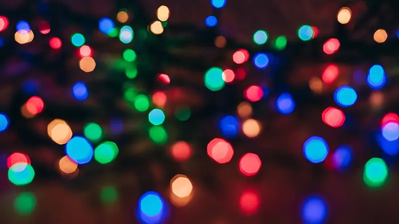 A blurred image of colorful holiday lights, including blue, green, red, and yellow bulbs, creating a festive and cheerful atmosphere. The lights appear to be intertwined and glowing softly against a dark background—capturing the essence of the best things to do on the Monterey Peninsula in November.
