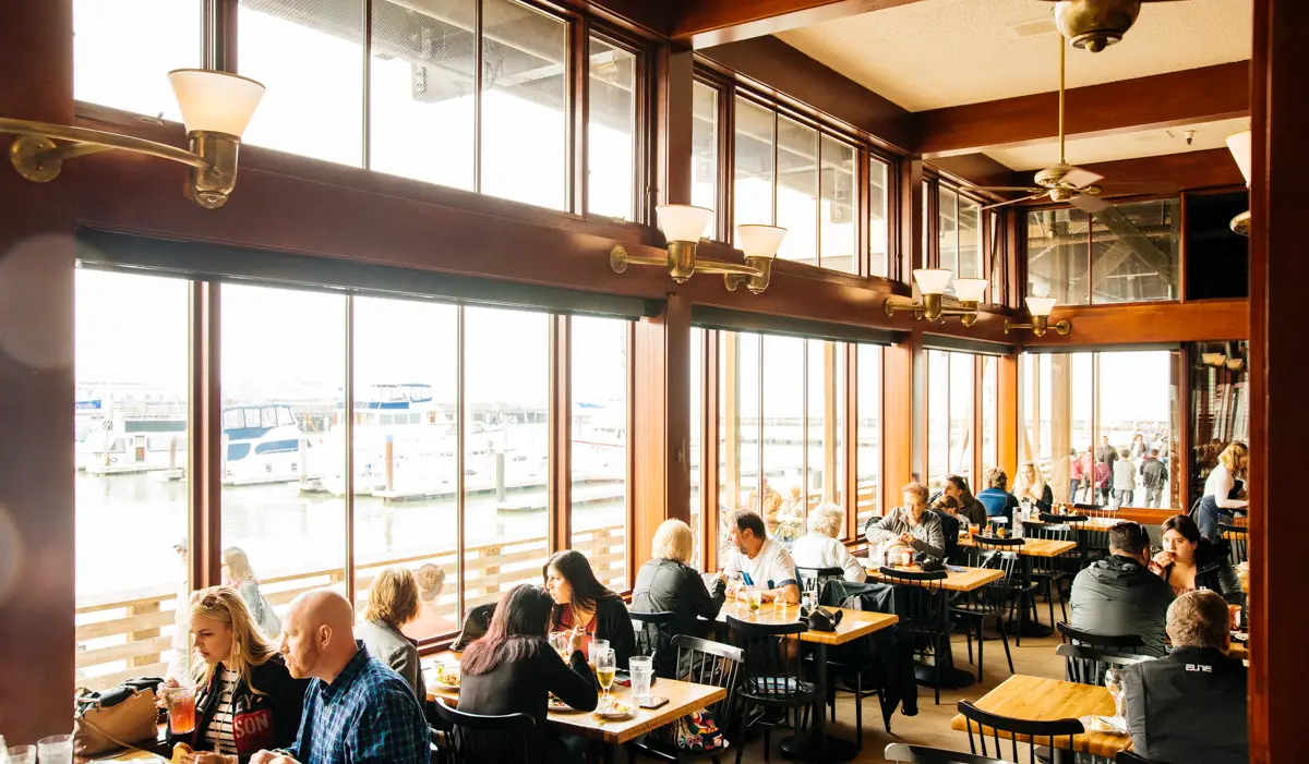 A bustling restaurant where to eat in the Bay Area, with large windows offering a view of boats docked at a marina. Diners are seated at wooden tables on the right, enjoying meals and conversations. The interior is warmly lit with modern light fixtures and has wooden accents.