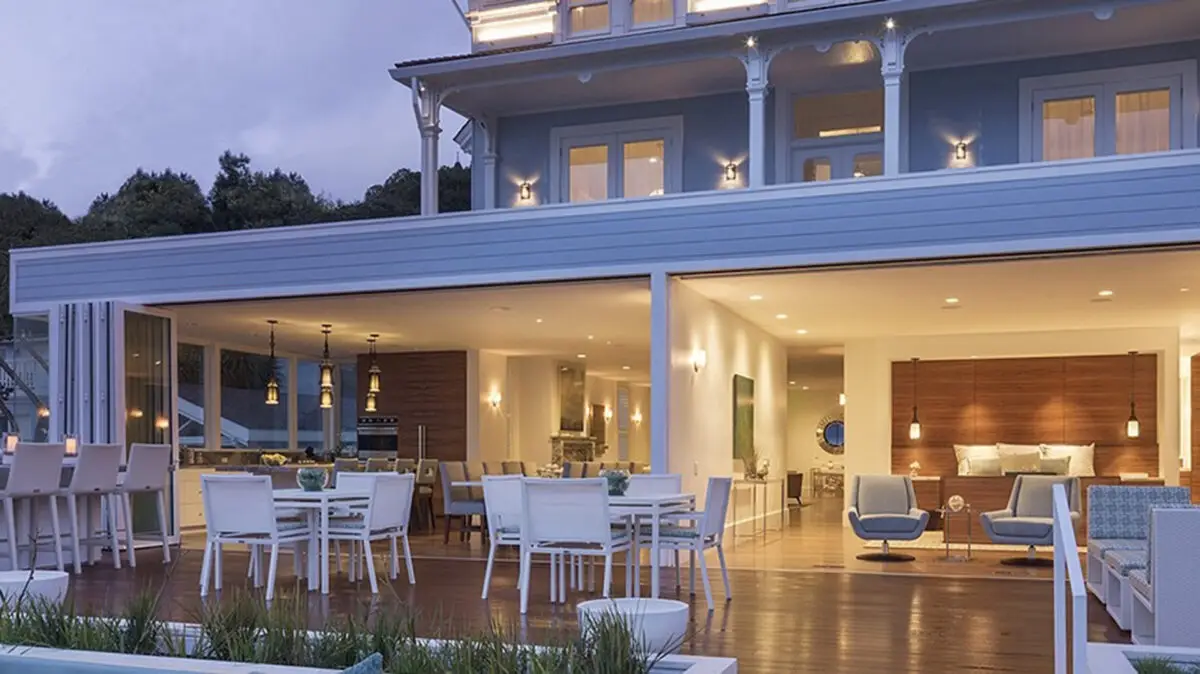 A modern two-story house with large, illuminated windows and an expansive outdoor deck. The deck features multiple white dining tables and chairs, resembling a chic spot from a "Where to Eat Bay Area" list. The indoor living area is visible through sliding glass doors, showcasing contemporary furniture and lighting.