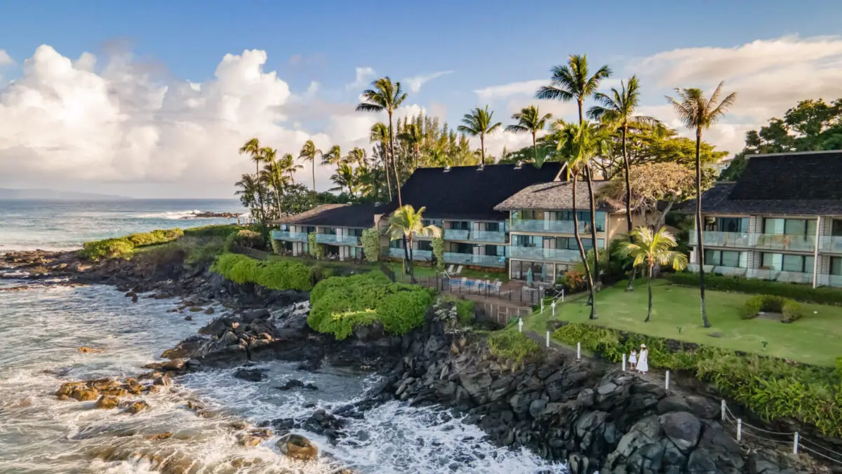 The Napili Kai Beach Resort is an oceanfront paradise nestled on a rocky coast, lined with palm trees and lush greenery. The multi-story buildings feature numerous balconies offering panoramic sea views, creating a true Hawaii home feeling as waves crash against the rocks below under a partly cloudy sky.