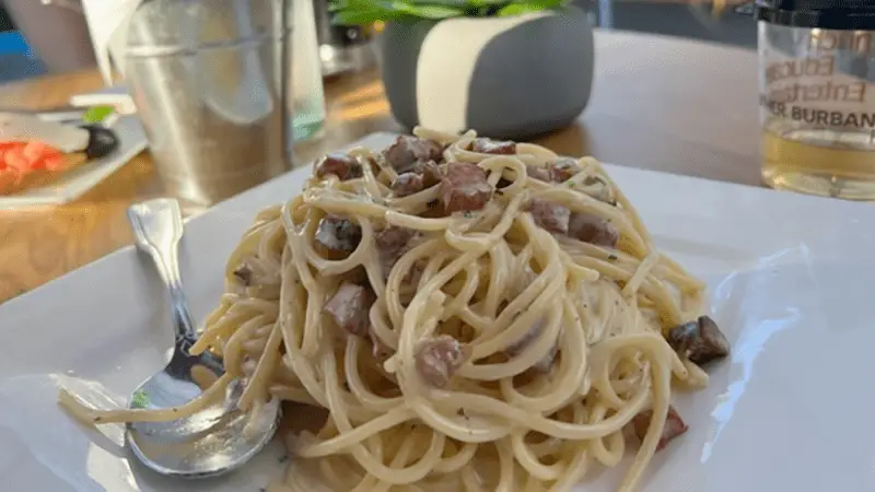 A white plate with a serving of creamy pasta mixed with diced meat pieces. A spoon is placed to the left of the pasta. The table, reminiscent of one you'd find at the best dinner spots in Sonoma County, also has a water glass, a salt shaker, and a potted plant in the background.