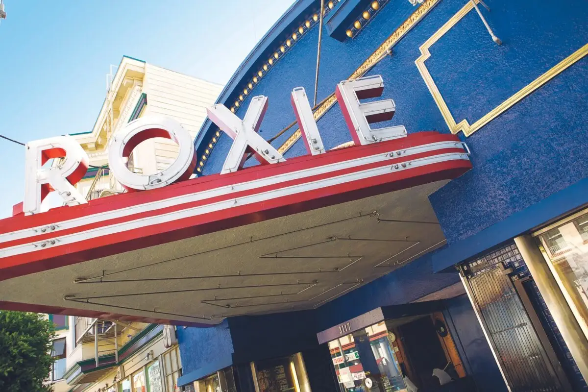 A blue theater building with a red-and-white marquee reading "Roxie." The marquee extends over the entrance and features large, illuminated letters. This iconic theater, one of the best things to do in San Francisco May, is located on a street with adjacent buildings visible in the background.