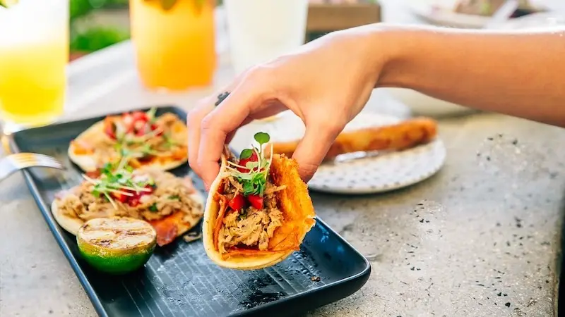 A hand is picking up a taco from a black rectangular plate, perfect for the best happy hour Waikiki has to offer. The taco is filled with shredded meat, microgreens, and red peppers. There is a grilled lime and other tacos on the plate. The table also has a drink and a small plate in the background.