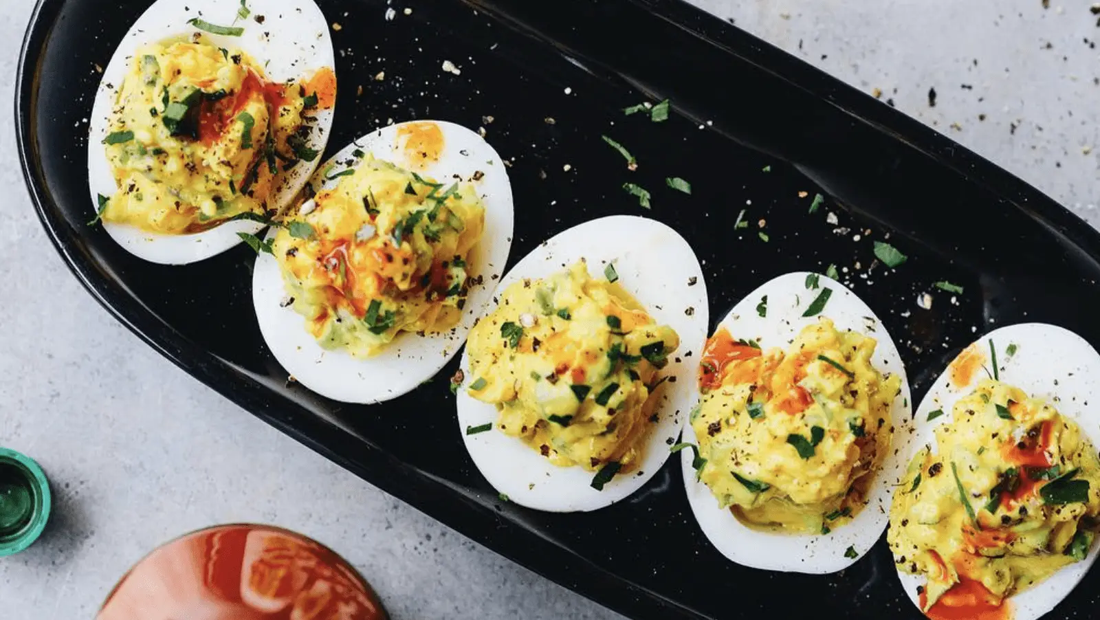 A black oval plate containing five deviled eggs, garnished with chopped herbs and a drizzle of hot sauce, is placed on a gray surface. A partly visible bottle of hot sauce is seen in the lower-left corner, making it the best lunch Napa Valley has to offer.