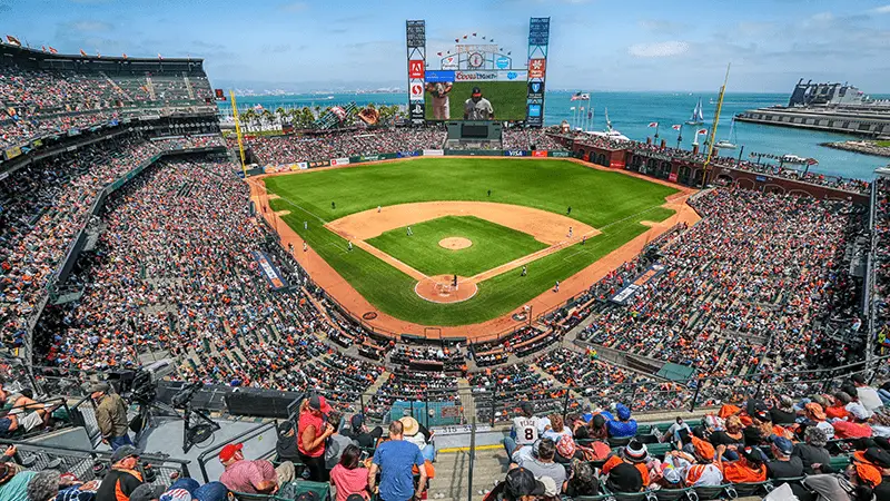 An aerial view of a packed baseball stadium with a game in progress, one of the best things to do in San Francisco this May. The field is surrounded by thousands of spectators, and a large scoreboard is visible in the background. Beyond the stadium, a body of water and a boat can be seen, with the city skyline in the distance.