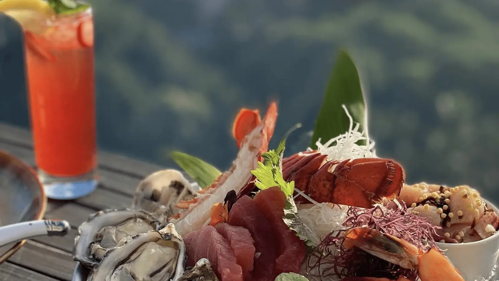 A seafood platter featuring oysters, tuna, shrimp, and other seafood delicacies is set on an outdoor table. A vibrant red cocktail with a lemon slice and straw is placed beside it. The background shows a blurred view of greenery and mountains, capturing the best sunset dining Oahu has to offer.