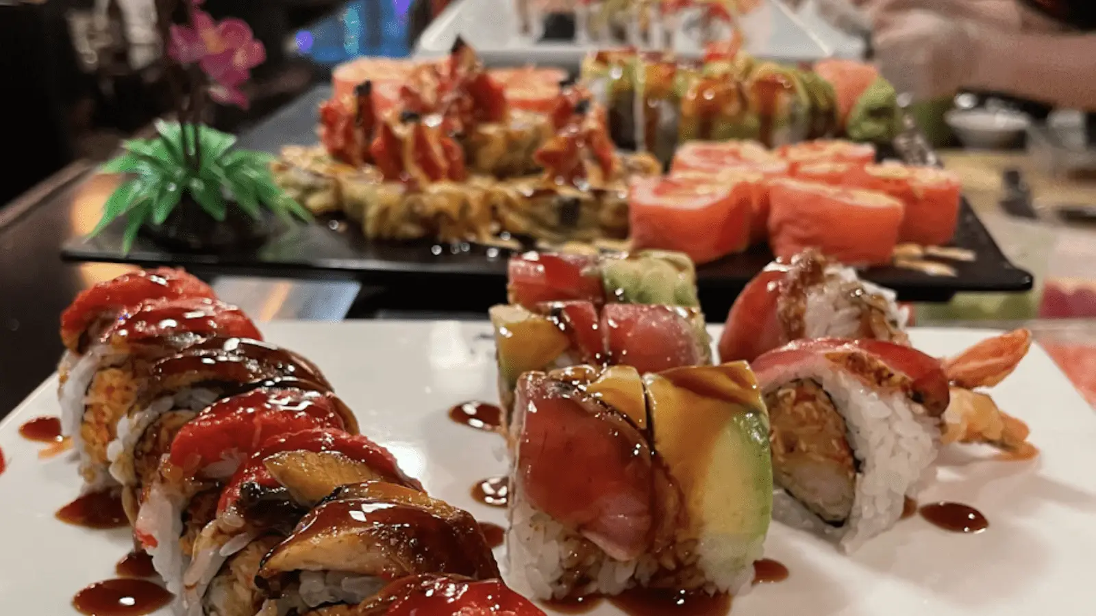 Close-up of assorted sushi rolls on plates. Various types of sushi, topped with colorful ingredients like avocado, sesame seeds, and different sauces, are artistically arranged. In the background, a chef's hands are visible preparing more dishes at the best sushi on the Big Island. A plastic decorative plant is also in view.
