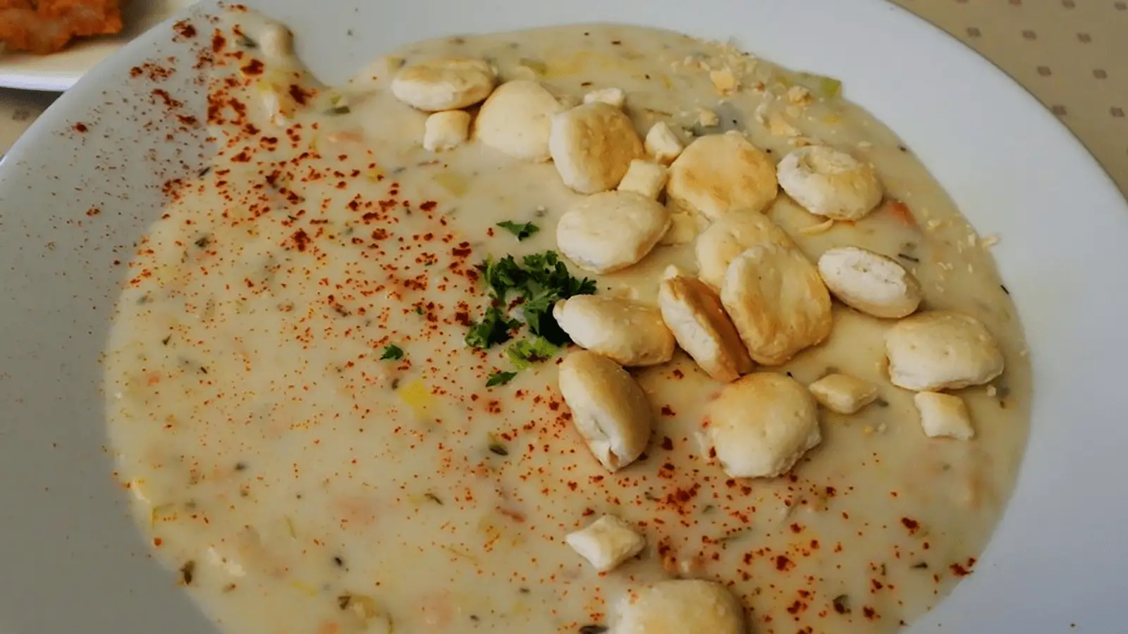 A creamy bowl of the best clam chowder Monterey Peninsula has to offer, topped with oyster crackers and sprinkled with parsley and red pepper flakes, served on a white plate.