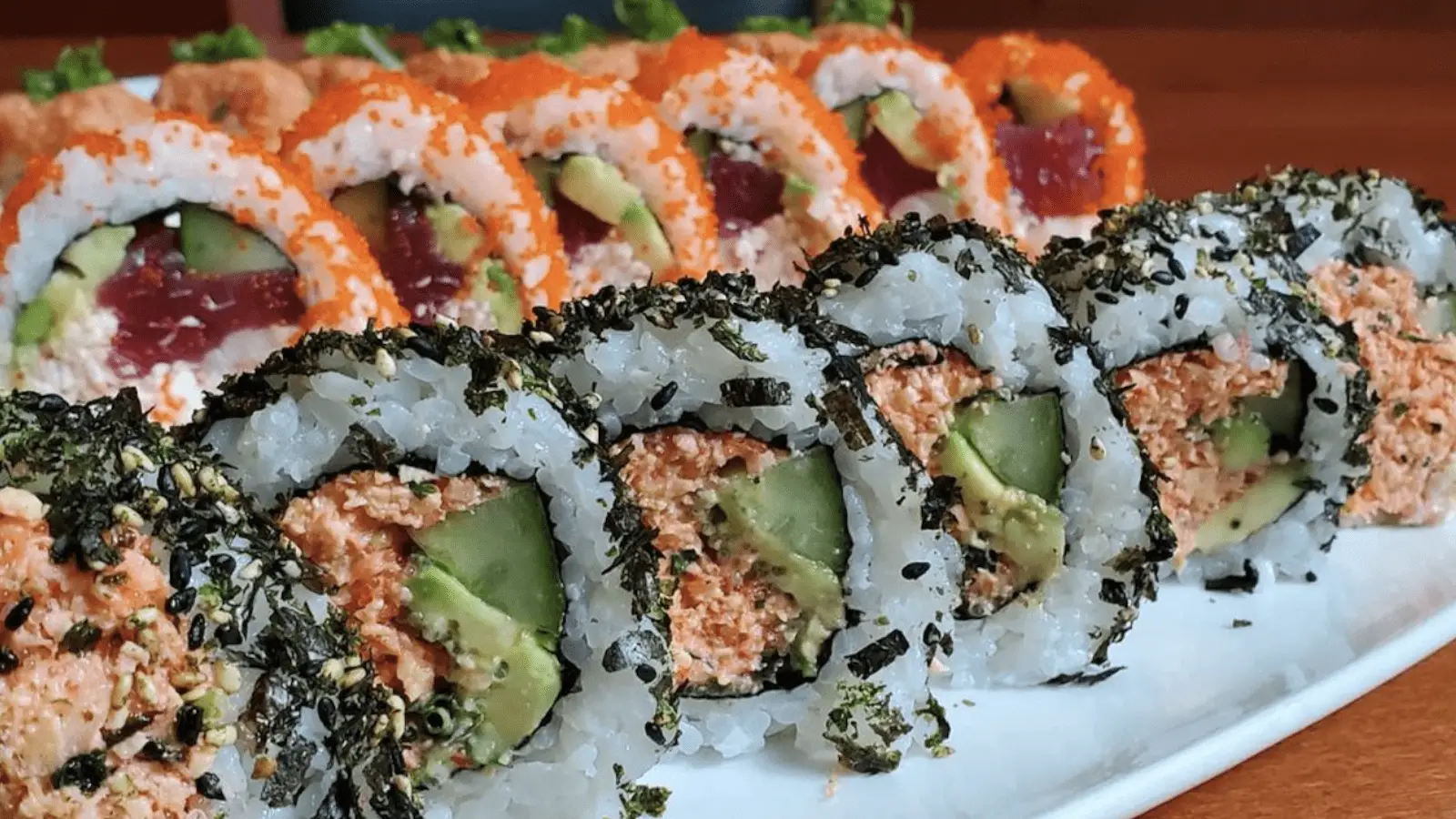 A plate of sushi rolls is displayed, reminiscent of the flavors found in Kohala and Waikoloa. The front row features rolls covered in nori and sesame seeds, filled with a mixture of avocado and what appears to be spicy tuna. The background row consists of rolls with orange roe on the outside, also containing avocado and tuna.